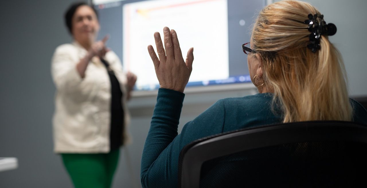 A student raising her hand in a computer lab, with instructor blurred in background